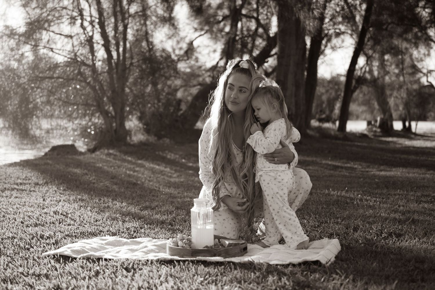 Black and white photo of a mother kneeling on a blanket in a park, hugging her toddler child outdoors.