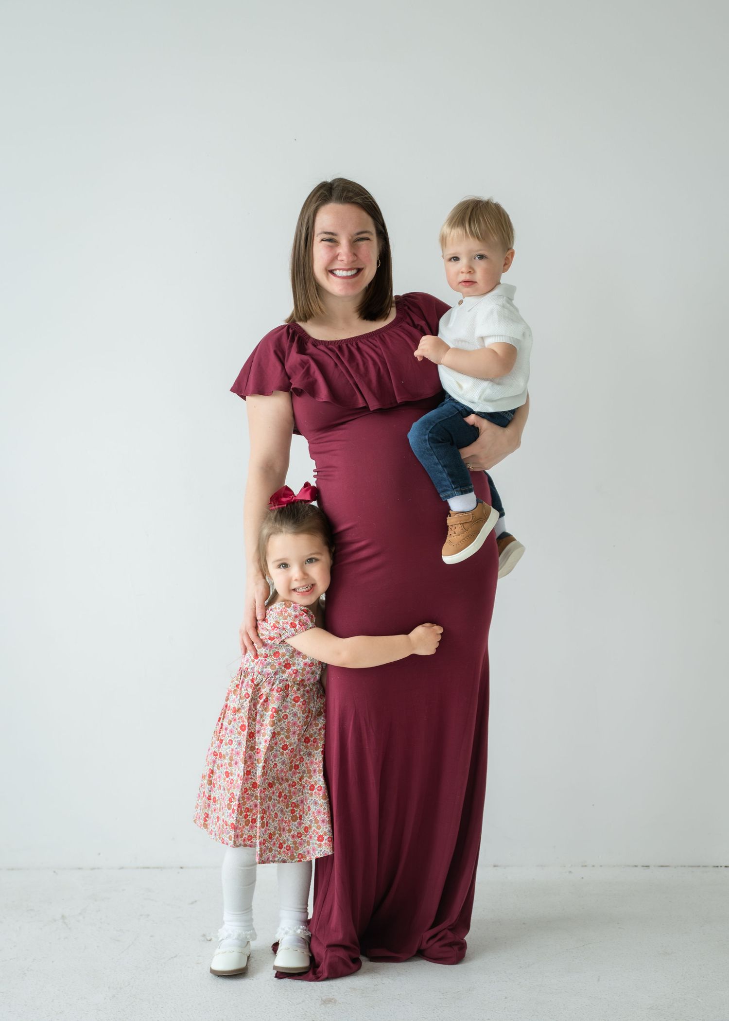 Pregnant woman in burgundy maxi dress smiling with toddler girl in floral dress and baby boy in white studio photo.