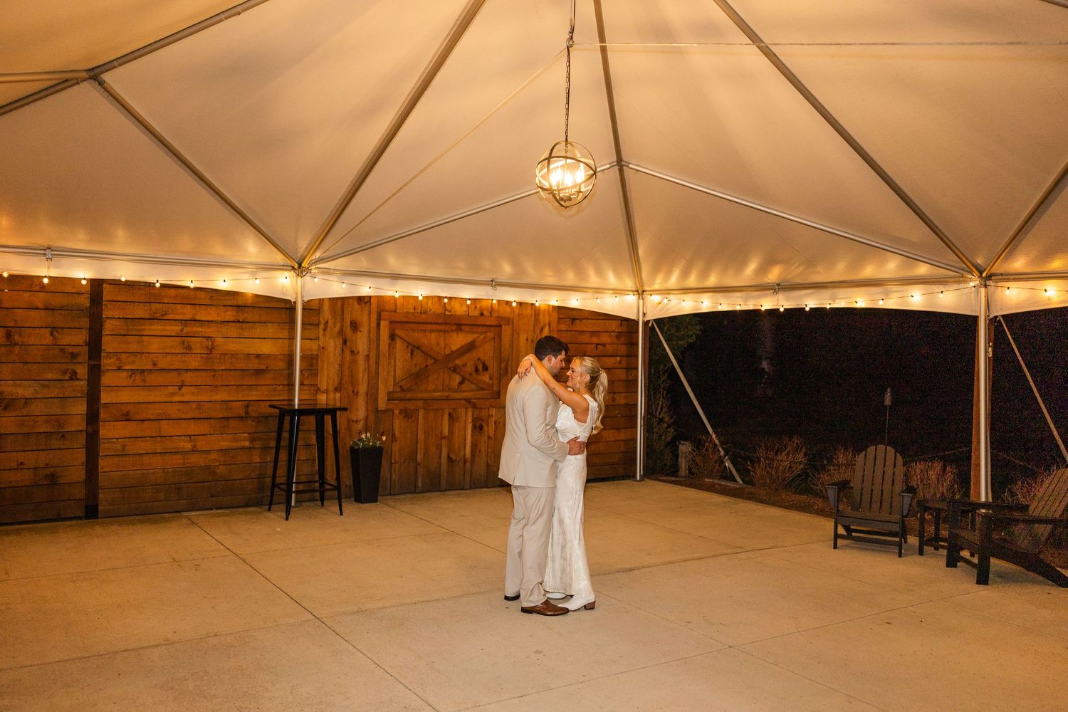 Couple sharing first dance under elegant white tent with warm lighting at rustic barn wedding venue.