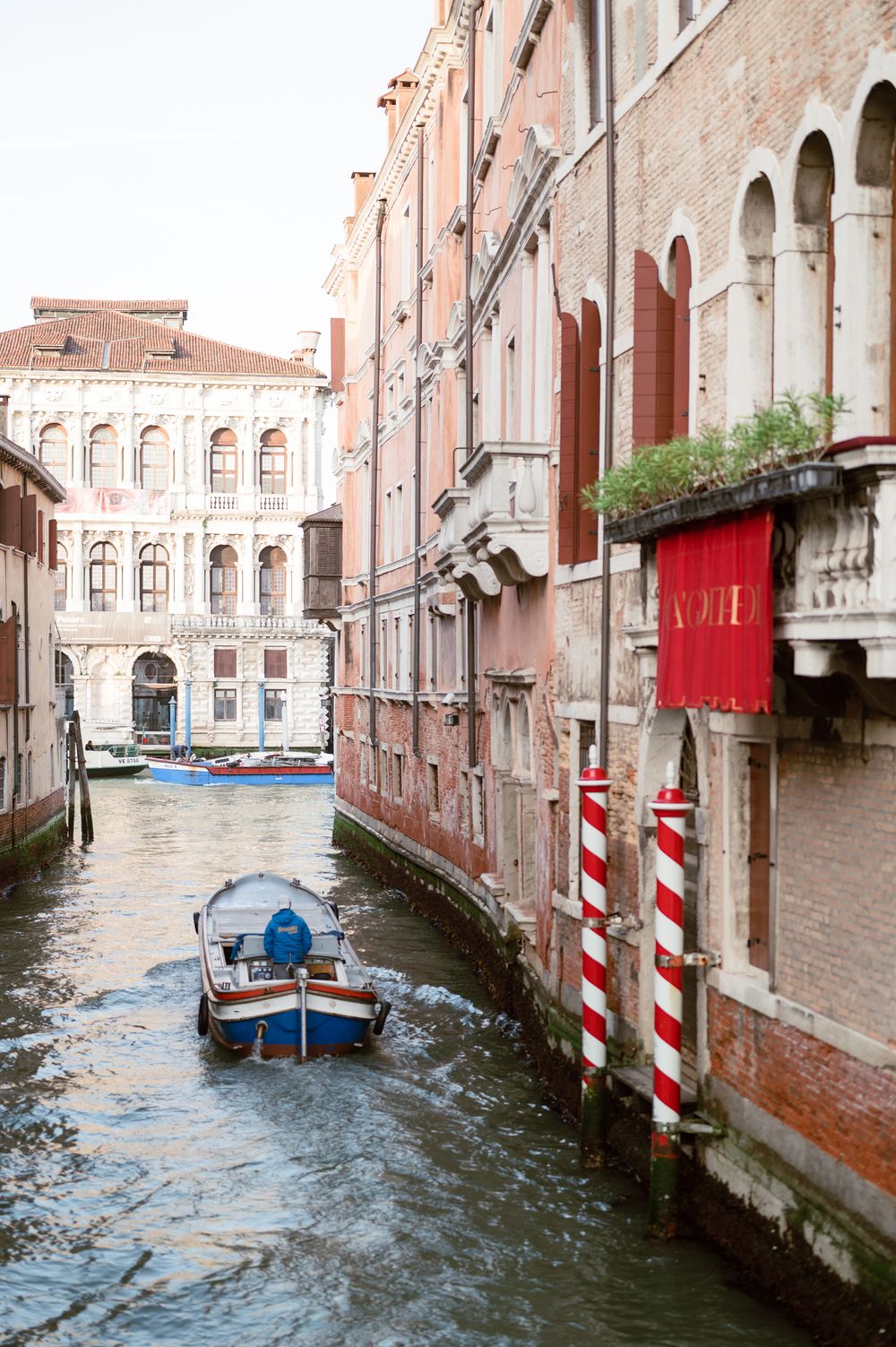A small boat navigates a narrow Venice canal flanked by historic pink buildings with arched windows and red shutters.