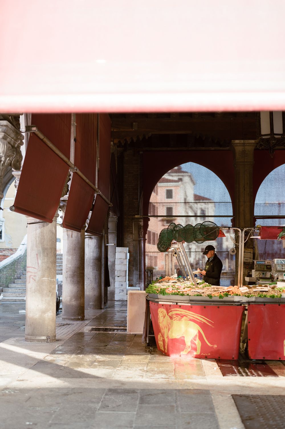Vendor at a vibrant red market stall under arched colonnade in Venice, Italy, arranging fresh produce.