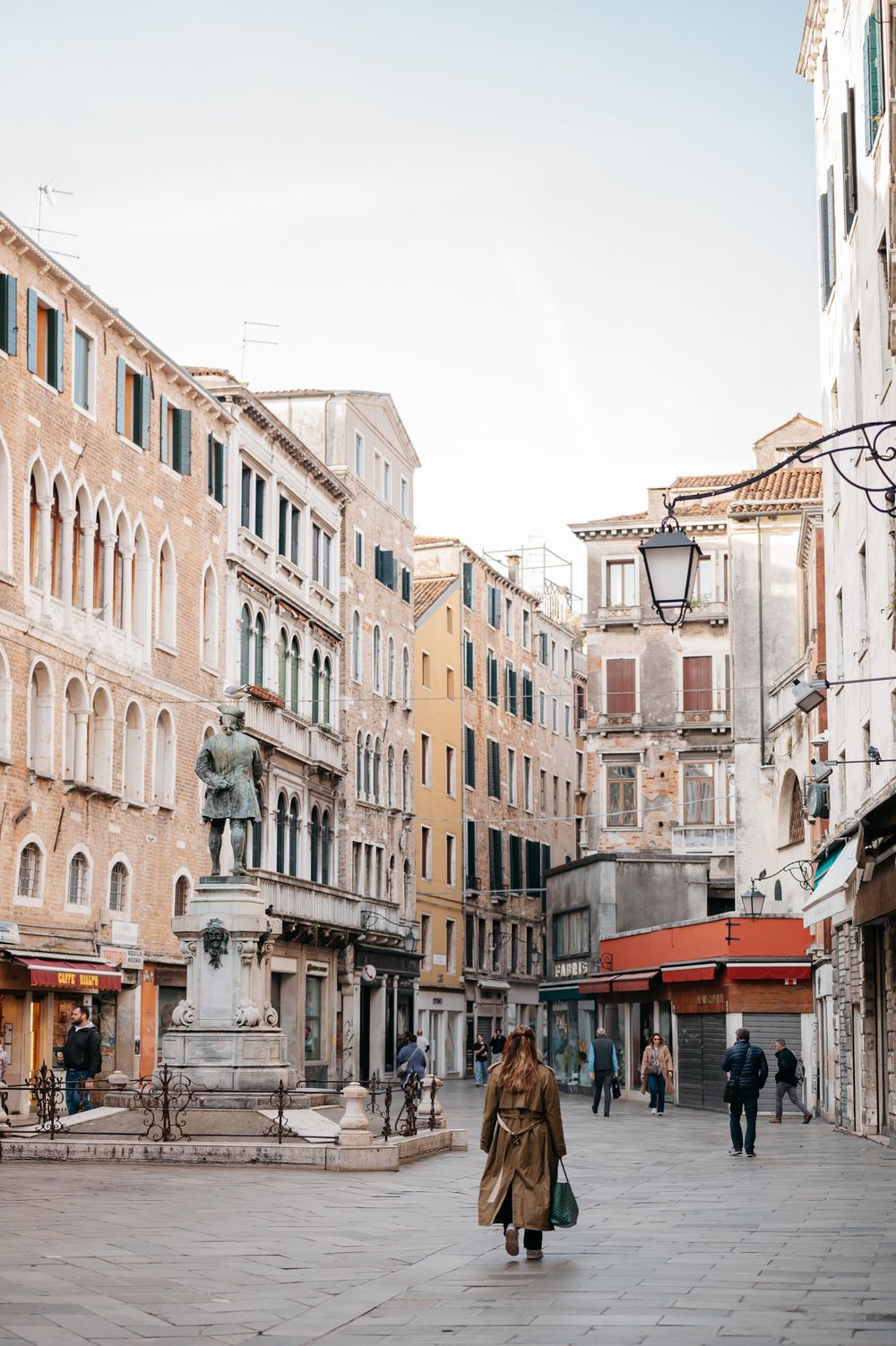 Woman in trench coat walking through a historic Venetian piazza with classical architecture and a bronze statue.