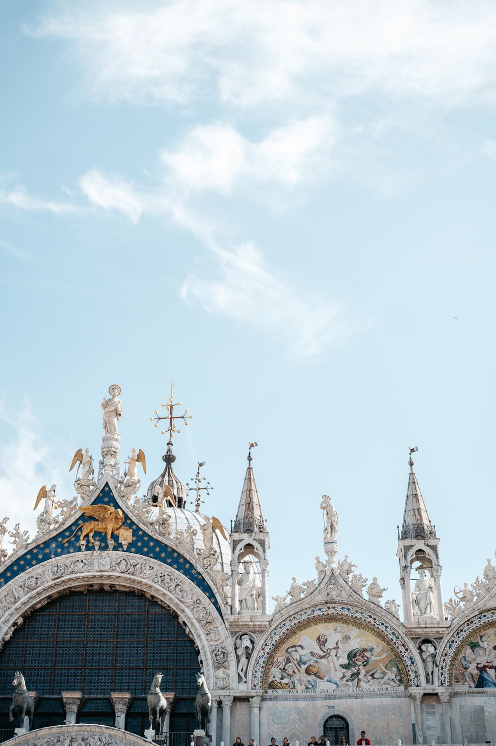 Ornate facade of St. Mark's Basilica in Venice, Italy, featuring golden mosaics, statues, and spires against a blue sky.