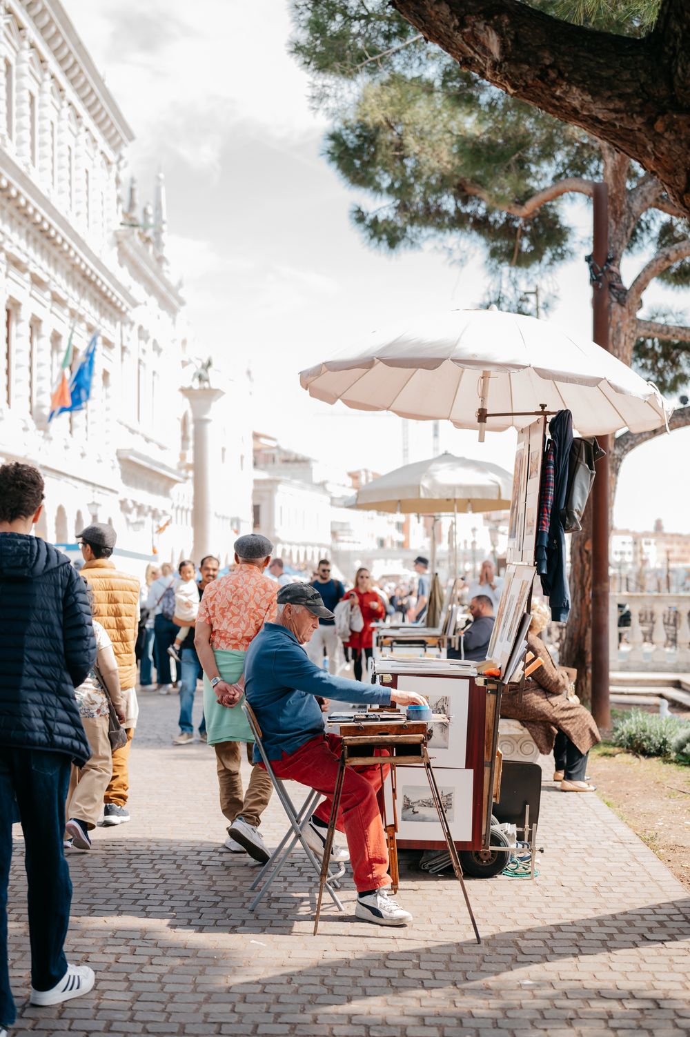 Artist painting outdoors on cobblestone street in Venice Italy with tourists walking nearby under umbrella.