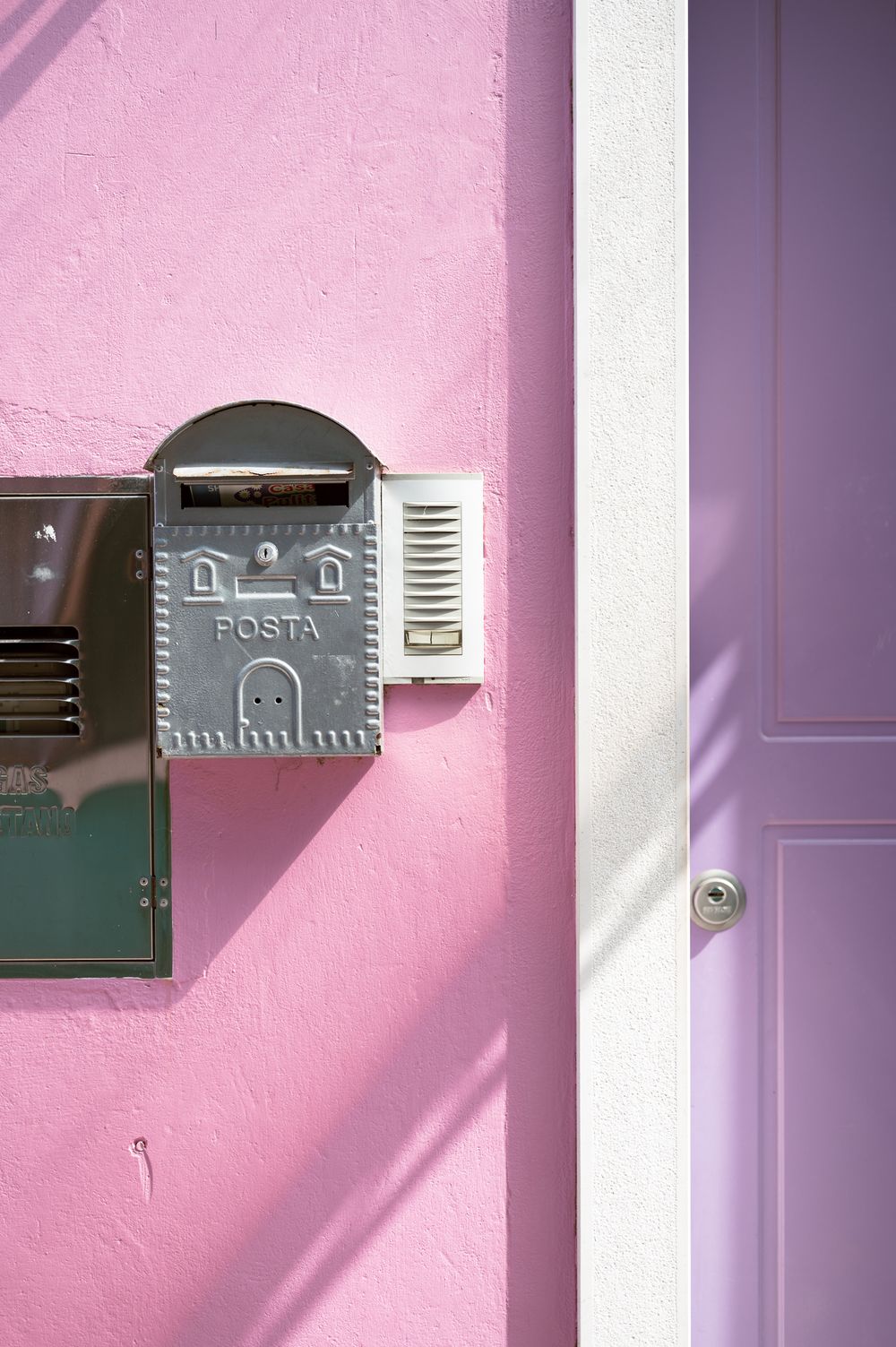 Silver POSTA mailbox and intercom mounted on a pink stucco wall beside a lavender door in sunlight.