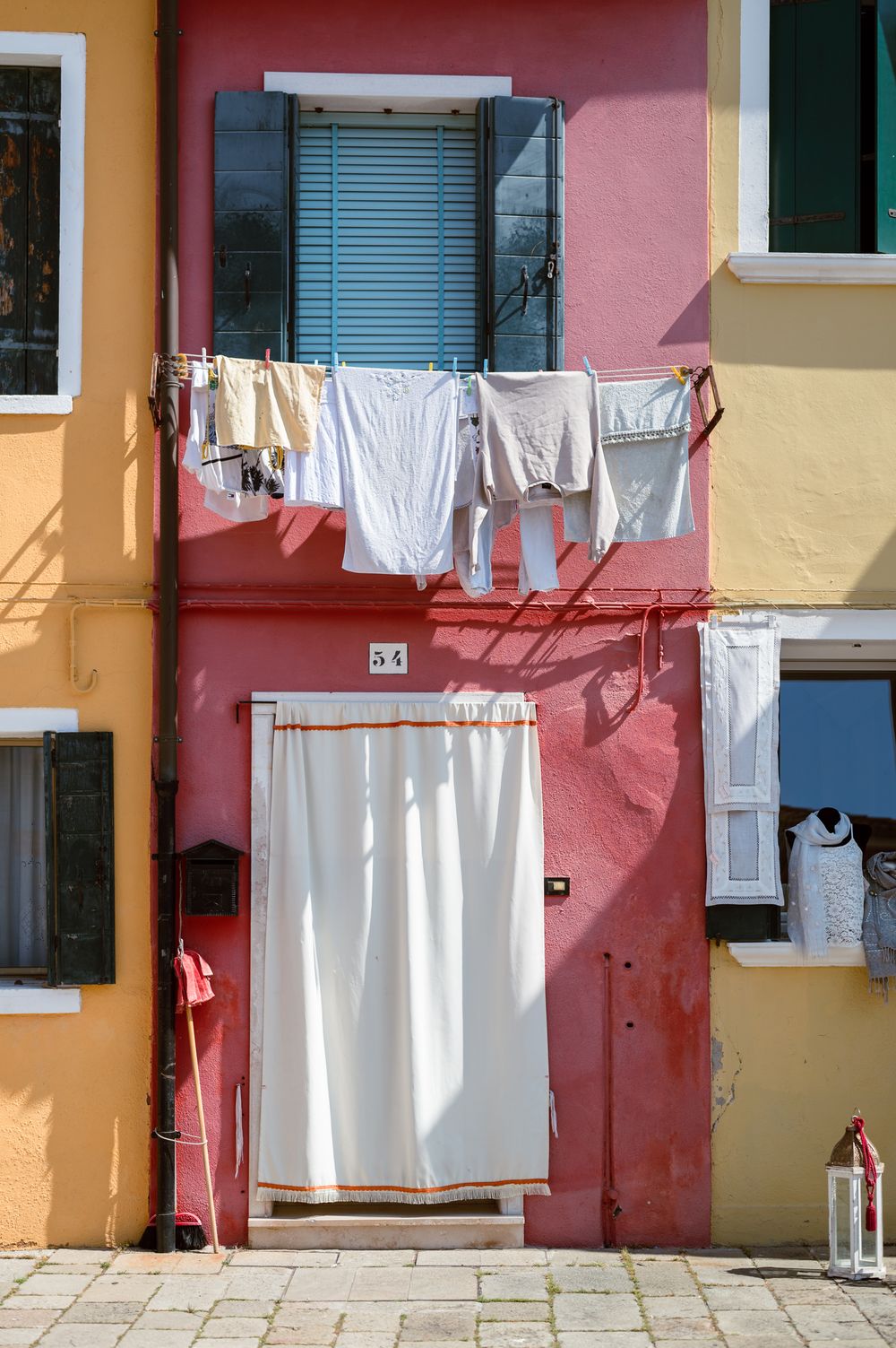 Colorful Burano building facade with laundry hanging on clothesline, red and yellow walls, blue shutters, white curtained door.