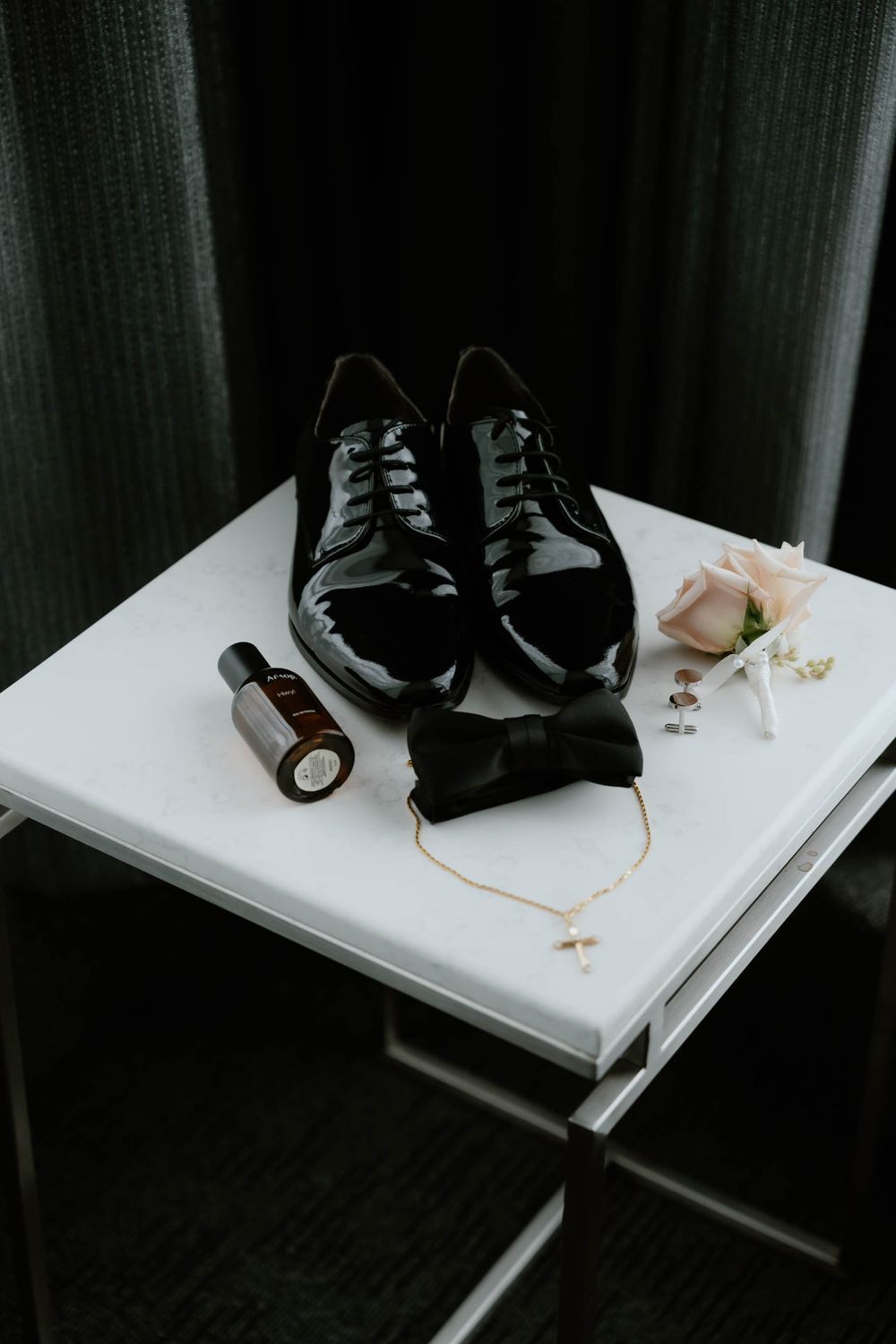 Black patent leather dress shoes, bow tie, gold cross necklace, boutonniere, and cologne arranged on a white table for a wedding.