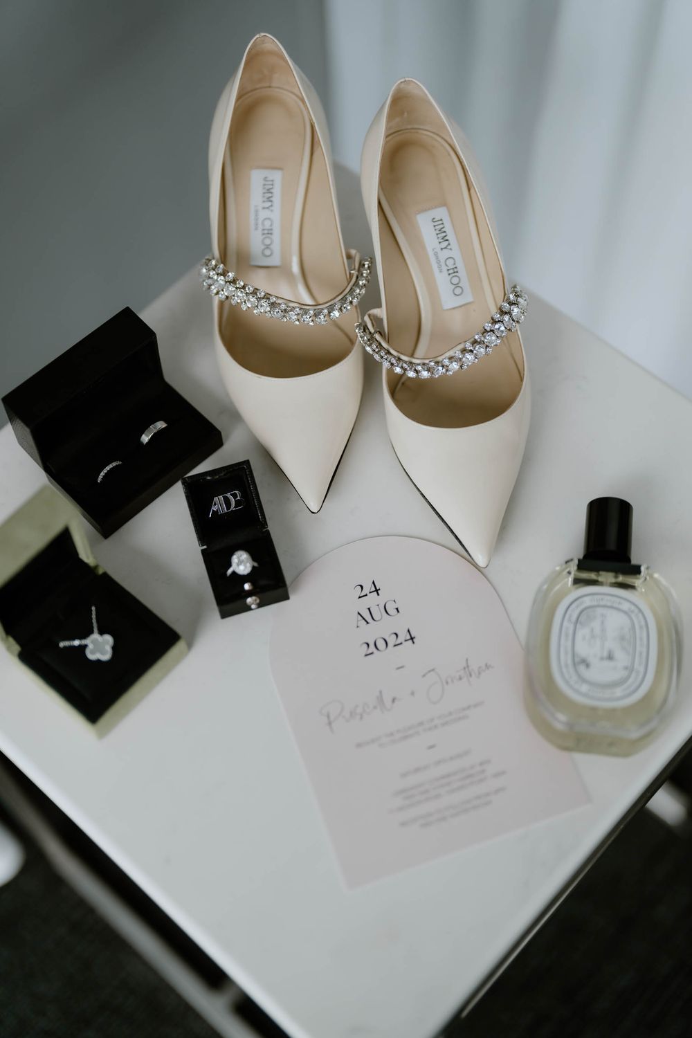 Elegant wedding flat lay featuring Jimmy Choo heels, diamond jewelry, Diptyque perfume, and a wedding invitation dated August 24, 2024.