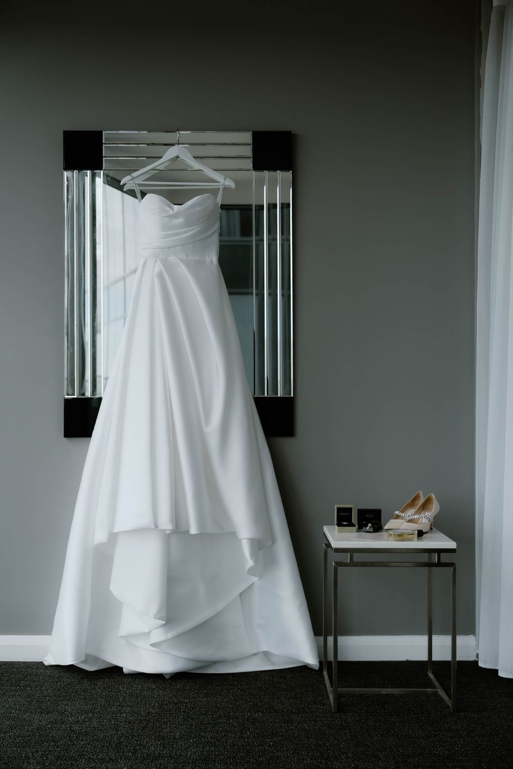 White strapless wedding gown hanging on mirror above small table with bridal heels and accessories in elegant hotel room.