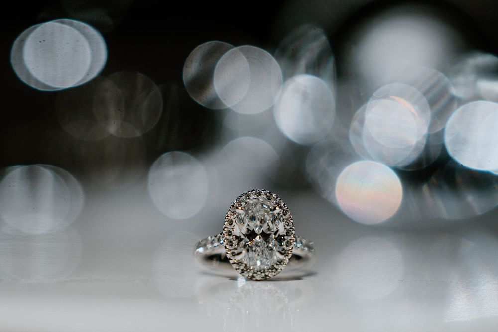 Elegant diamond halo engagement ring on white surface with soft bokeh lights in background.
