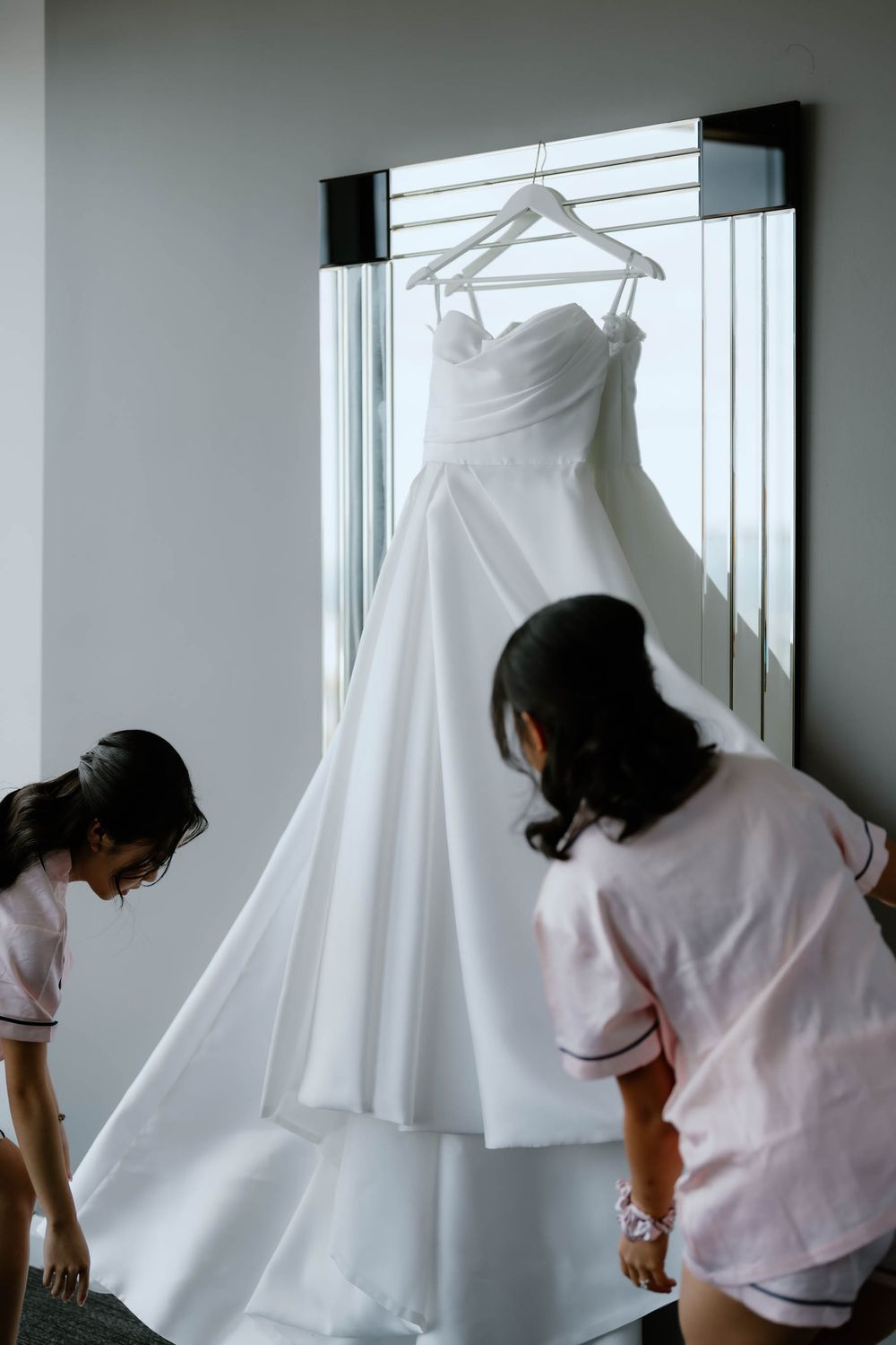 Two bridesmaids in pink robes admiring a white strapless wedding gown hanging on a mirror.