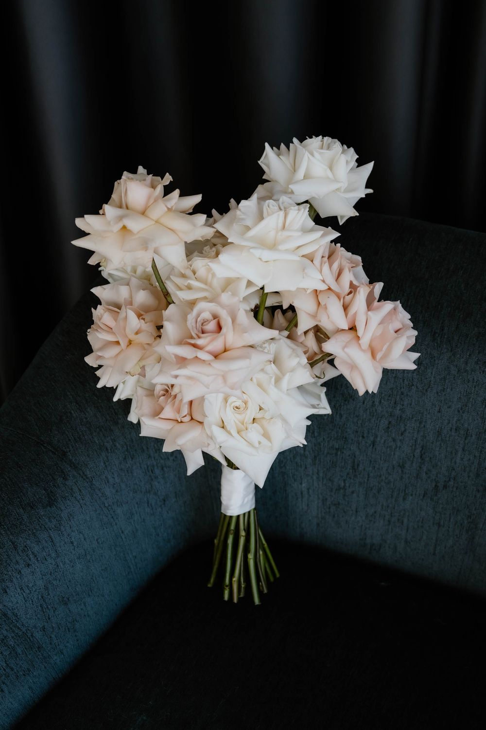 Elegant bridal bouquet of blush and white roses tied with ribbon resting on a dark velvet chair.