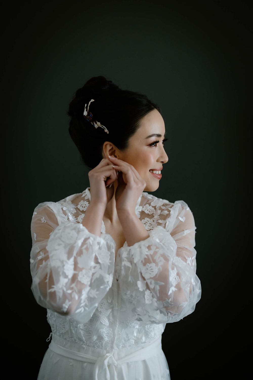 Smiling bride in white lace dress adjusting her earring, with elegant updo and hair accessory against dark background.