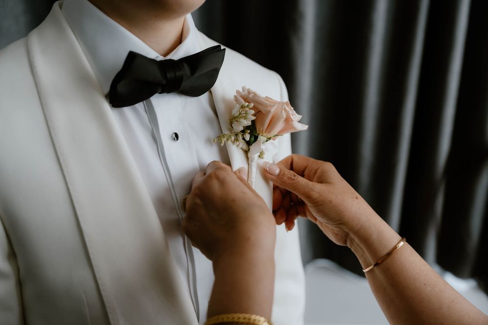 Hands pinning a white floral boutonniere on a groom's white tuxedo with black bow tie at a wedding.