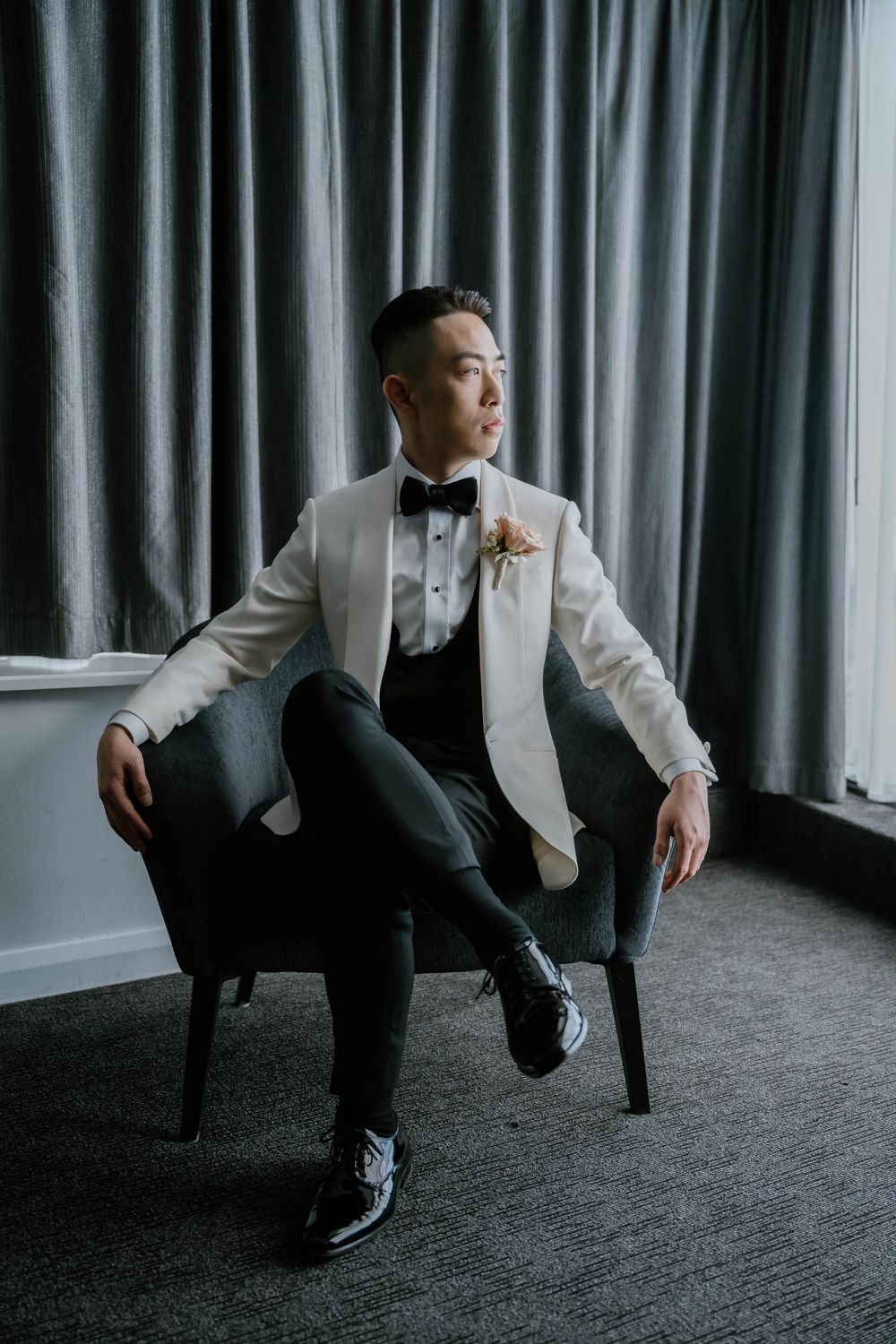 Dapper groom in white tuxedo jacket and black bow tie seated in velvet chair by curtained window.