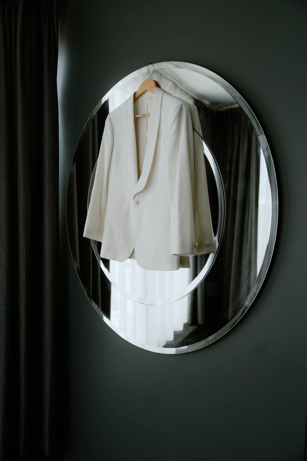White blazer on wooden hanger displayed in oval silver mirror against dark green wall.