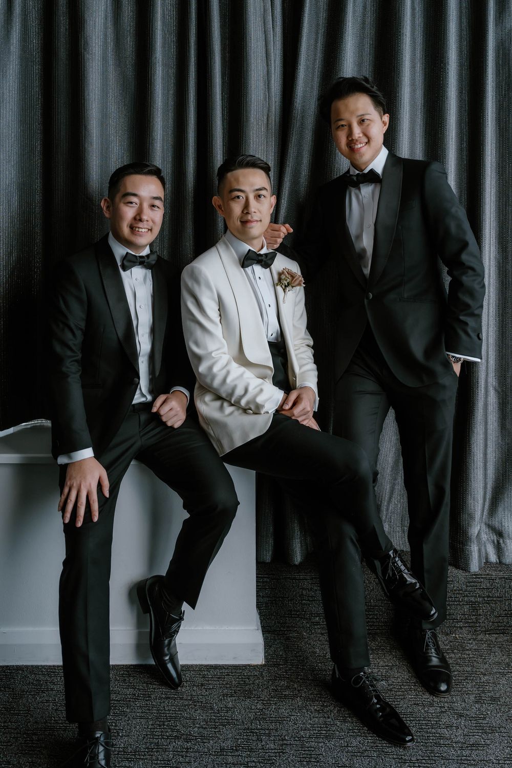Three groomsmen in black tuxedos and bow ties pose elegantly, with the groom in a white jacket and boutonniere.