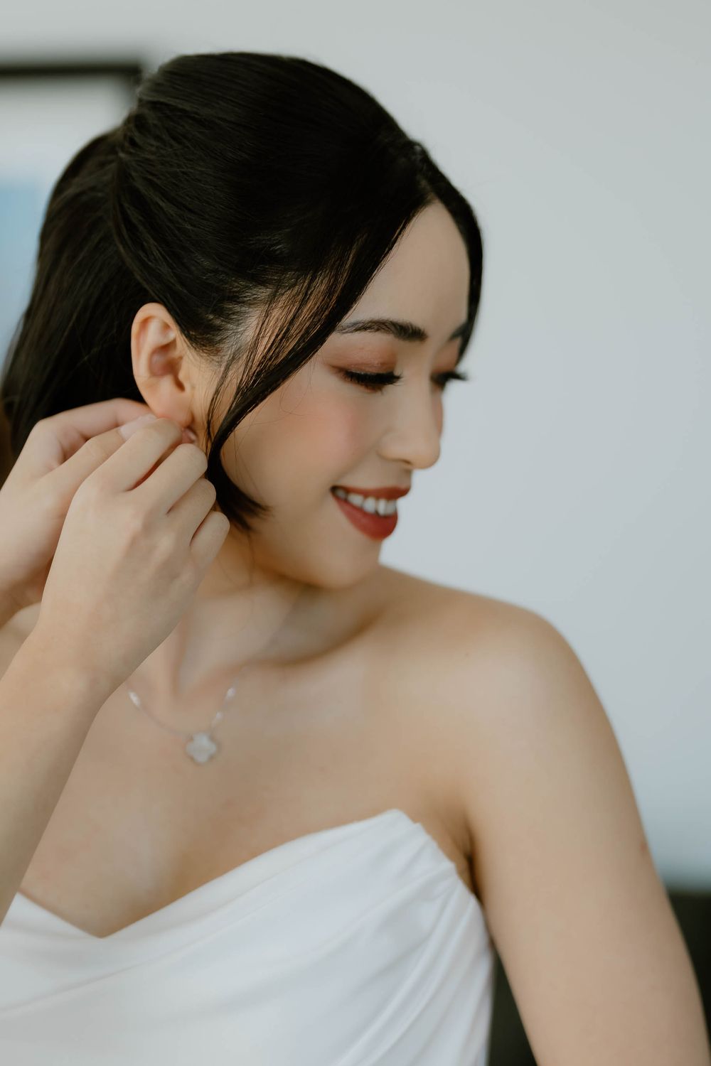 Beautiful Asian bride smiling while putting on earrings wearing strapless white gown and delicate necklace.