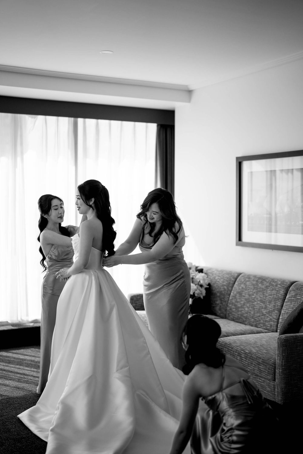 Black and white photo of bride being helped into wedding gown by bridesmaids in elegant hotel room.