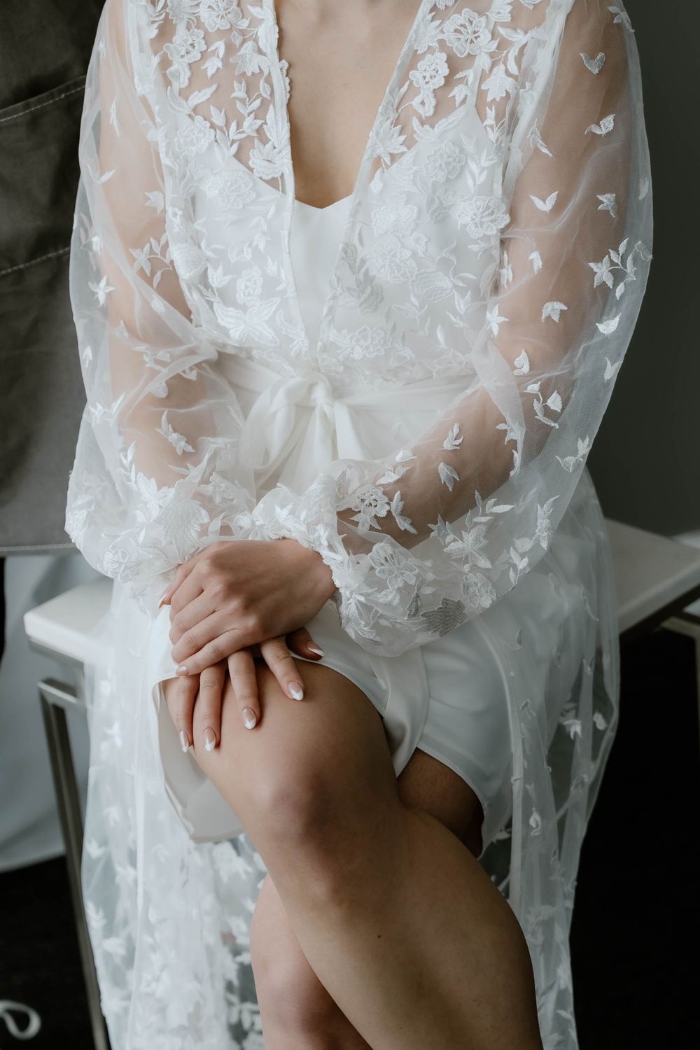Bride in sheer white lace robe with floral embroidery sits elegantly, hands folded gracefully on her lap.