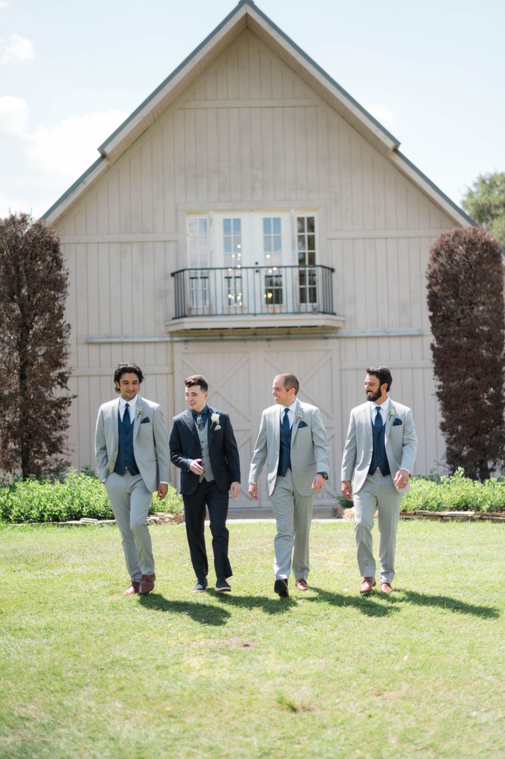Groom and groomsmen in gray suits walk outside rustic barn wedding venue on sunny day.