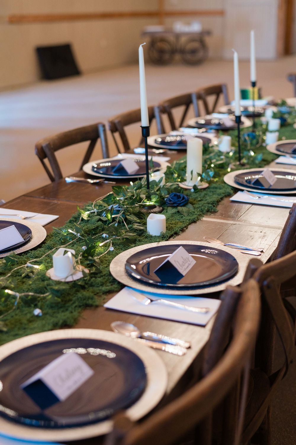 Elegant wedding reception table with green moss runner, navy charger plates, black taper candles, and fairy lights.