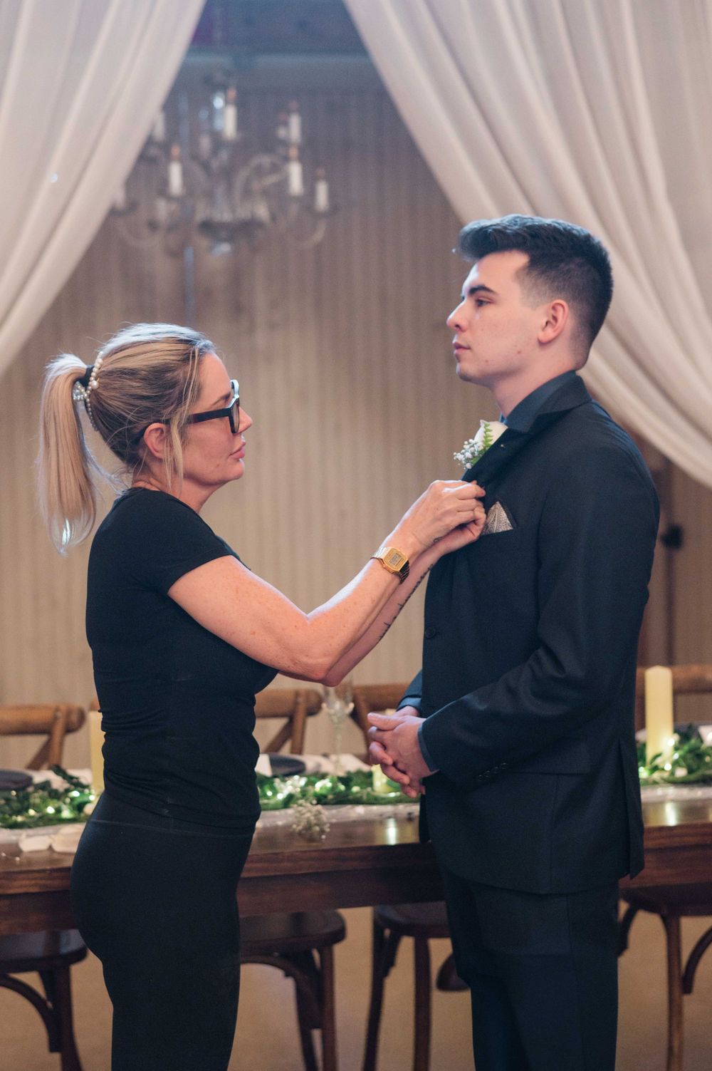 Wedding coordinator pins boutonniere on groom in black tuxedo at elegant venue with white drapes.