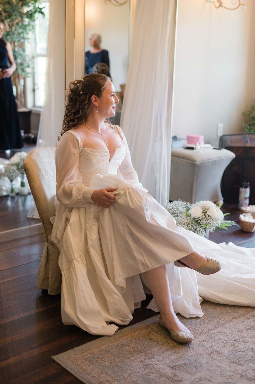 Bride in white gown with sheer sleeves sits gracefully in a chair, gazing out a window while getting ready.