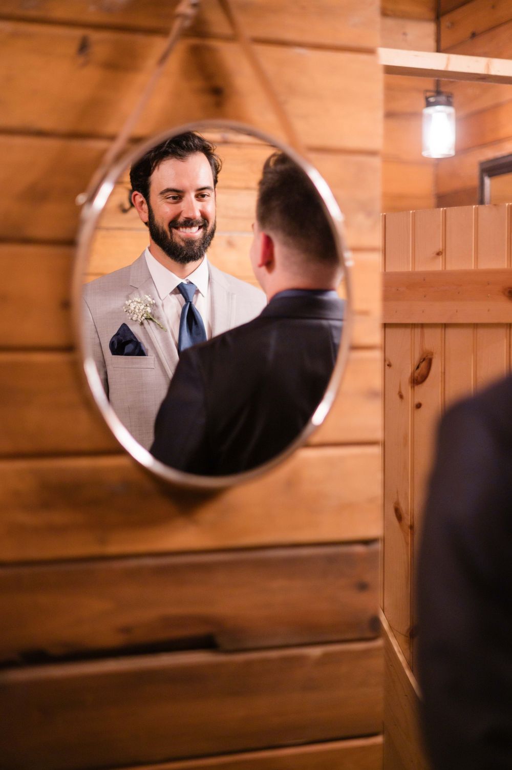 Smiling groom in gray suit with boutonniere reflected in round mirror inside rustic wooden cabin.