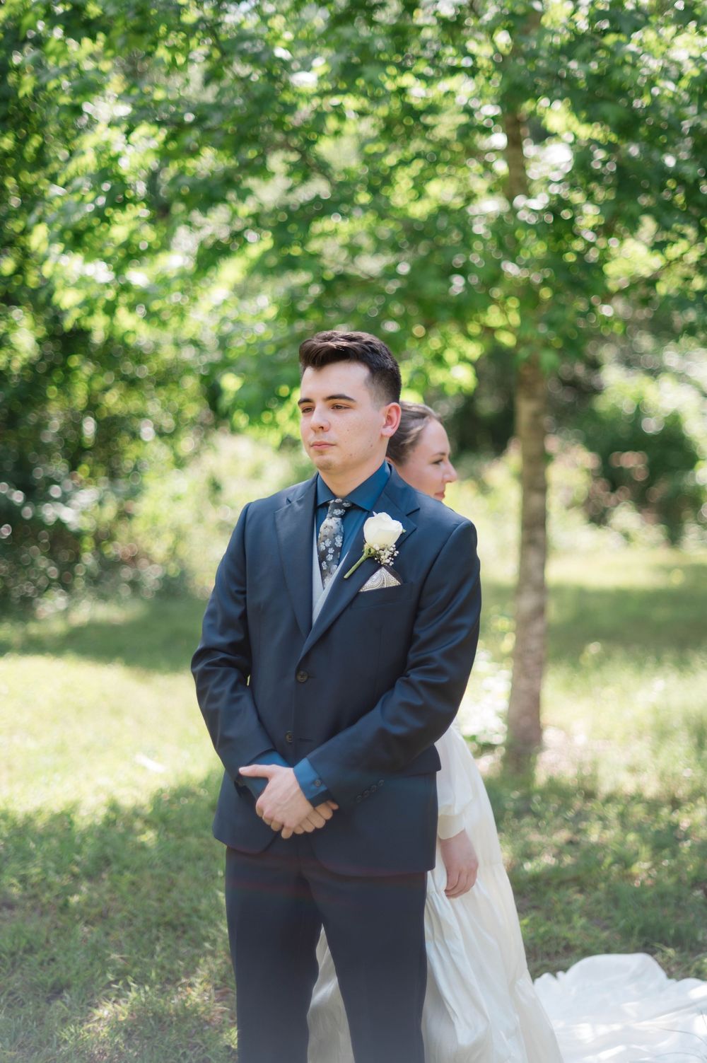 Groom in navy suit with white boutonniere stands outdoors as bride approaches in white gown during wedding ceremony.