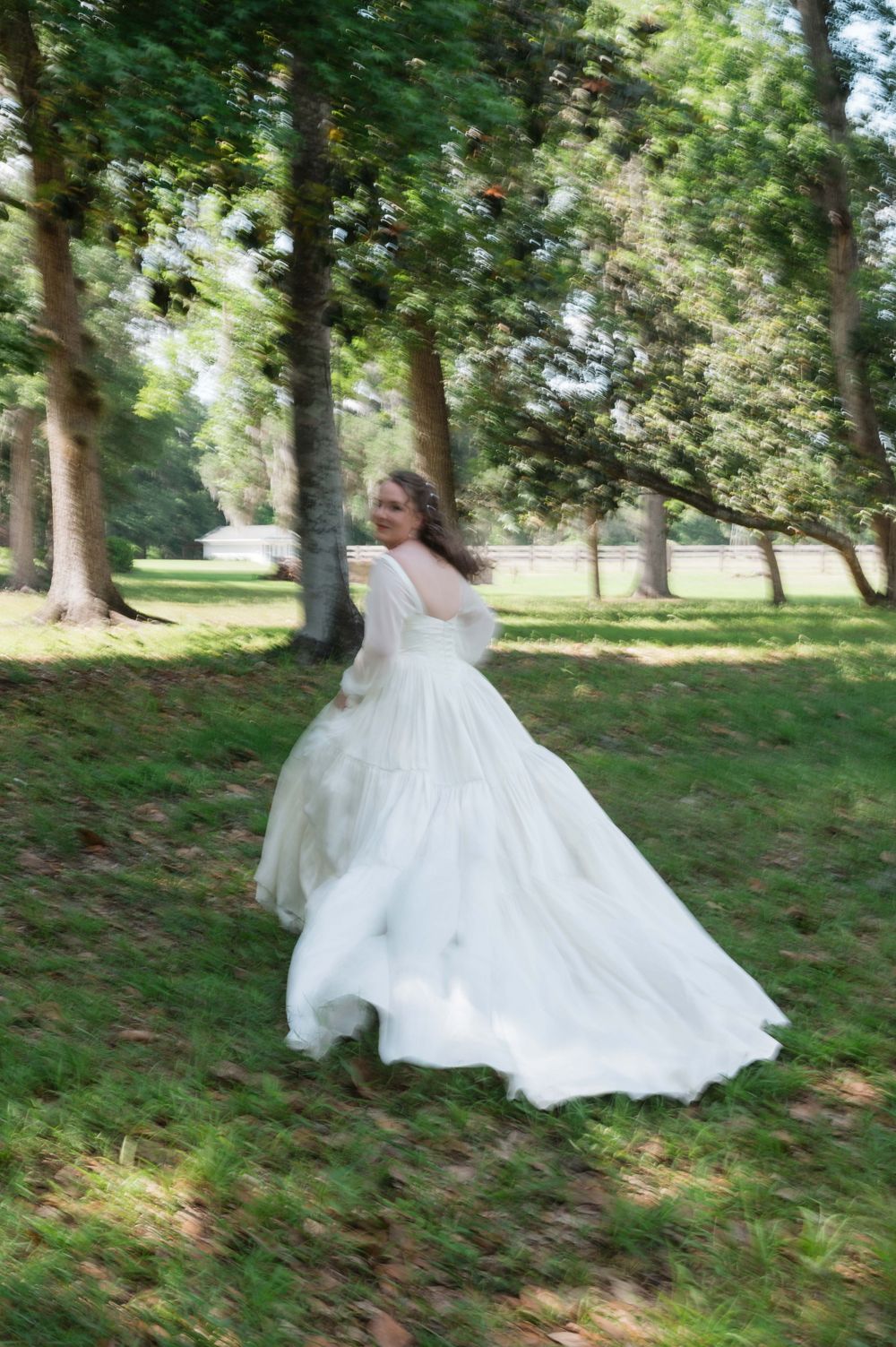 Bride twirling in flowing white wedding gown outdoors among green trees on lush grass.