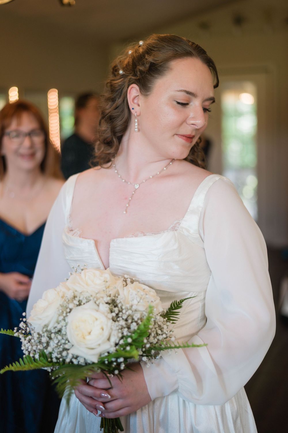 Bride in white gown holding white rose bouquet with baby's breath, wearing pearl jewelry at wedding ceremony.