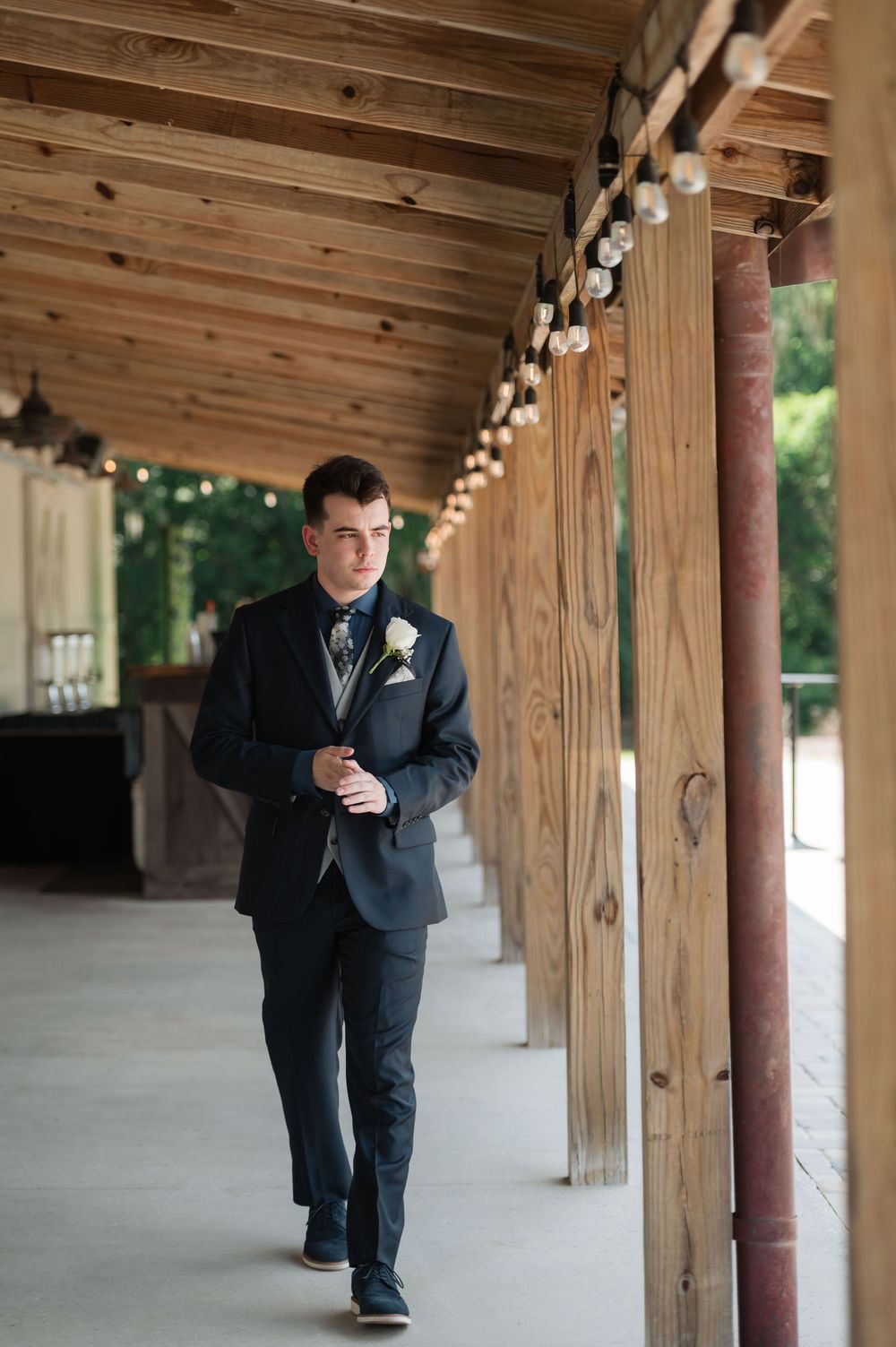 Dapper groom in navy suit with boutonniere walks confidently along a rustic wooden covered walkway with string lights.