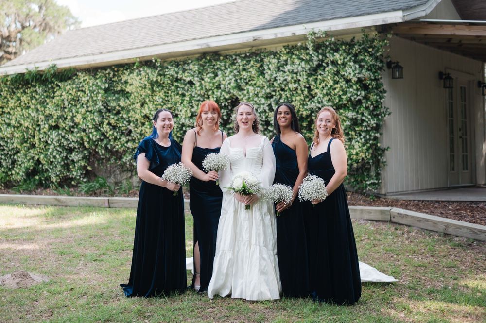Bride in white gown poses with four bridesmaids in navy blue dresses, holding bouquets in front of an ivy-covered building.