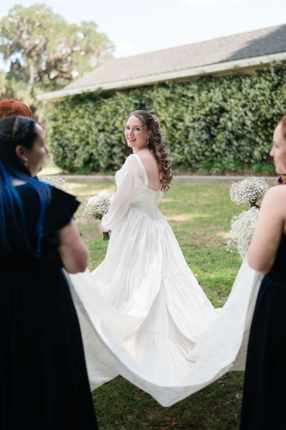 Bride in white gown smiles back as bridesmaids in navy dresses hold her train outdoors.