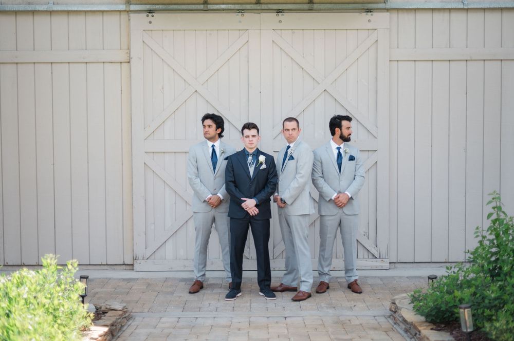 Four groomsmen in light gray suits with blue ties stand confidently in front of a rustic barn door at a wedding.