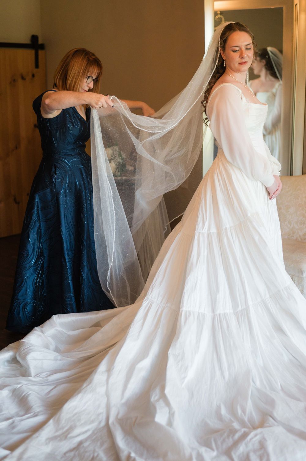 Mother helps bride in flowing white gown and long veil prepare for wedding ceremony indoors.