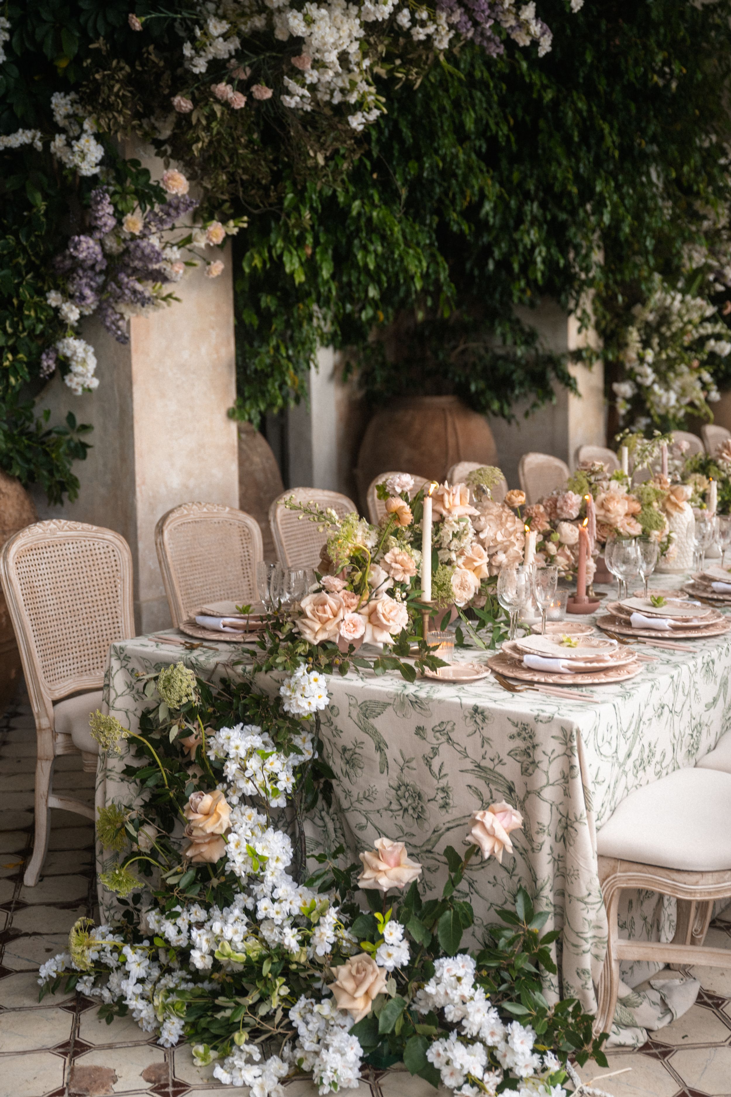 Elegant outdoor wedding reception table with floral tablecloth, peach roses, candles, and lush greenery backdrop.