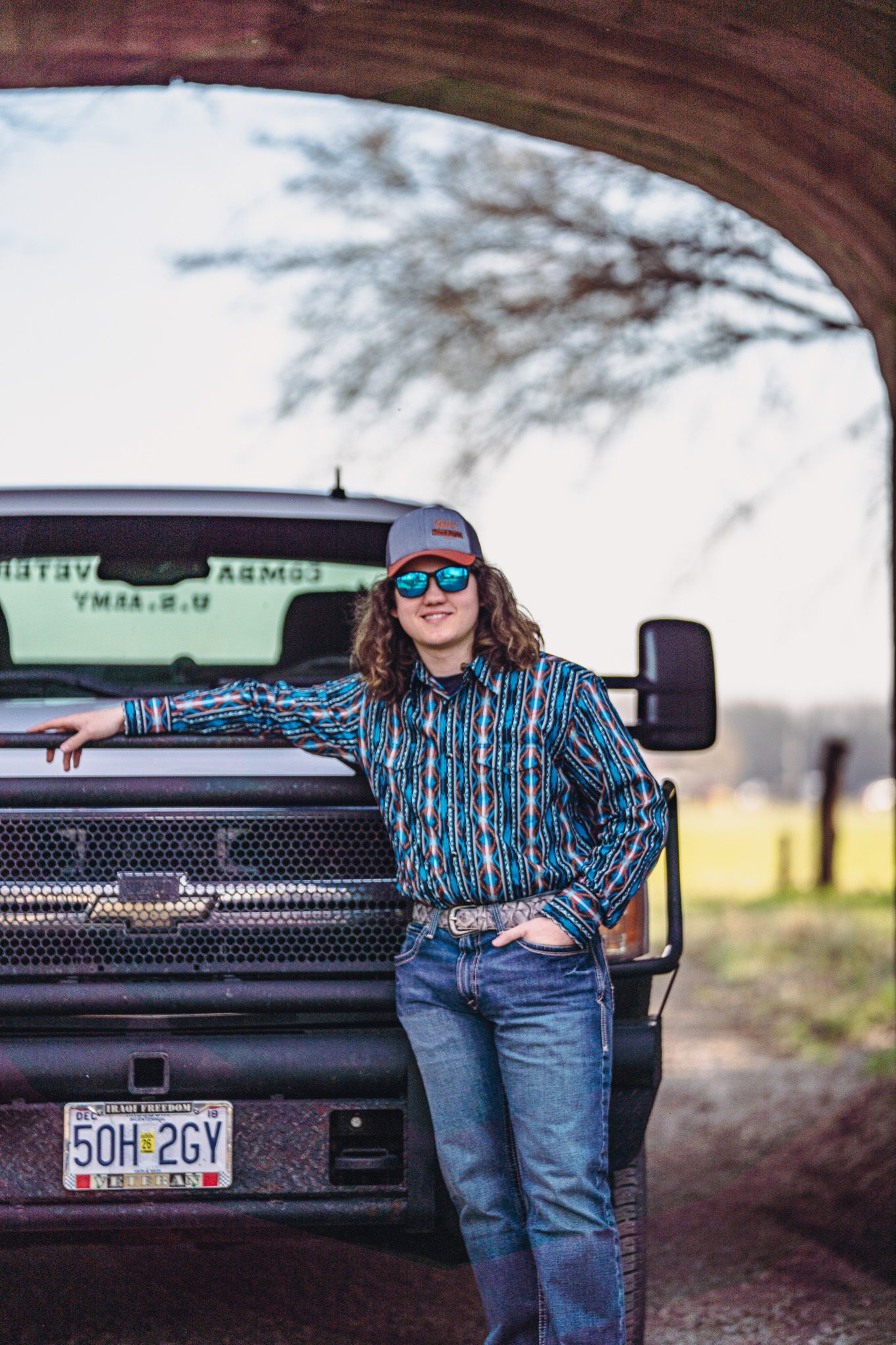 Young woman in western shirt and cap leans against a pickup truck on a rural road.