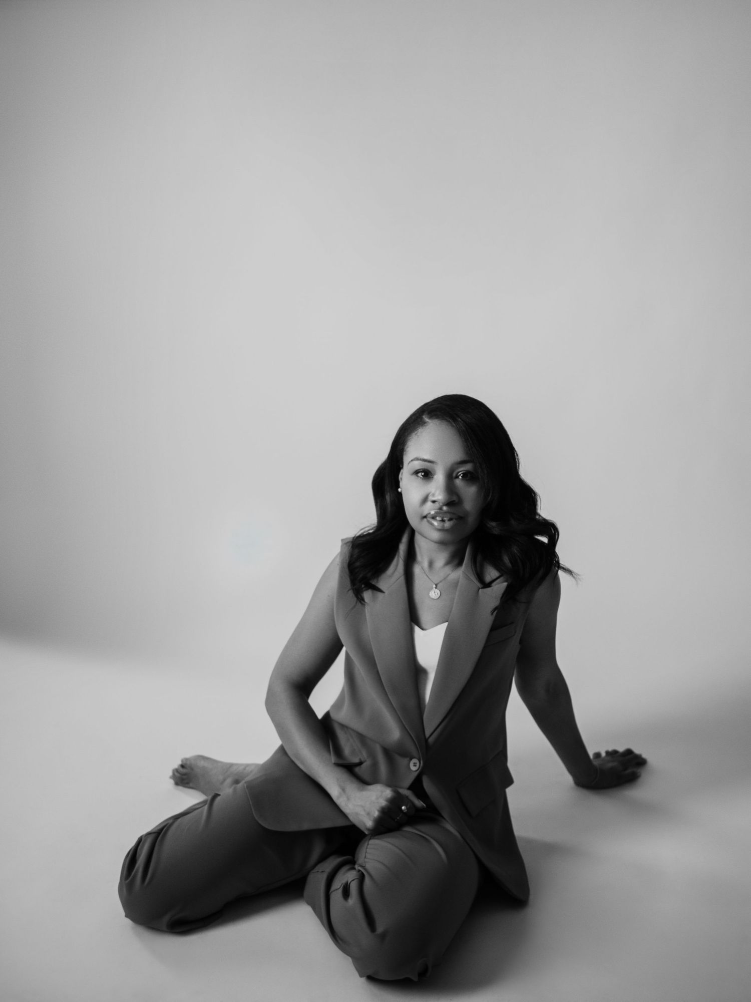 Black and white portrait of a woman in a blazer and trousers sitting gracefully on a studio floor.