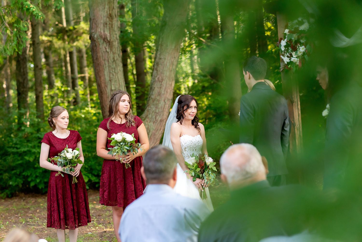 Bride and bridesmaids in burgundy dresses stand during an outdoor woodland wedding ceremony surrounded by lush green trees.