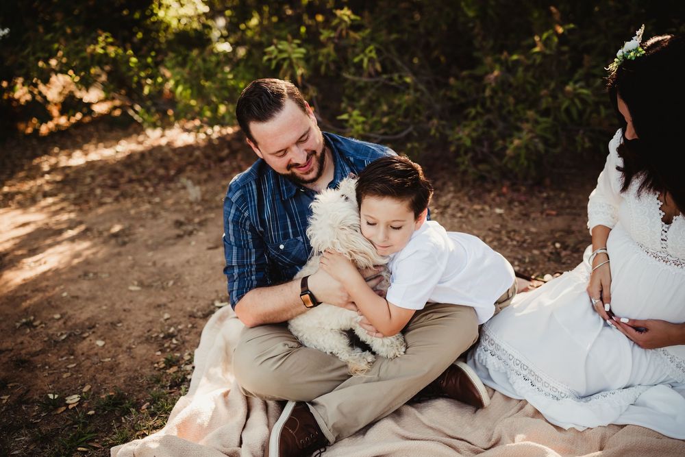 Family sitting on blanket outdoors, father and son laughing while holding white fluffy dog, pregnant mother beside them in redlands ca