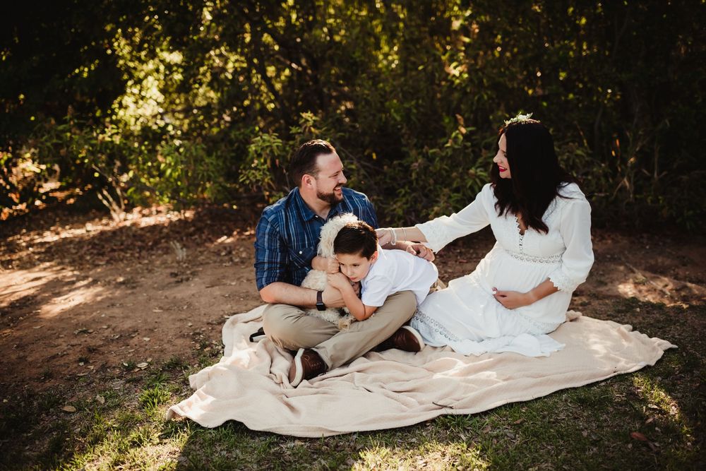 Pregnant woman in white dress sits on blanket outdoors with husband and child during family maternity photo session.