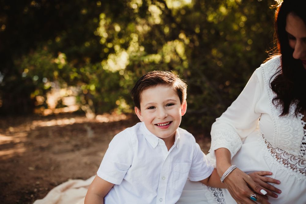Smiling boy in white shirt sits outdoors with a woman in white dress, surrounded by lush green trees.