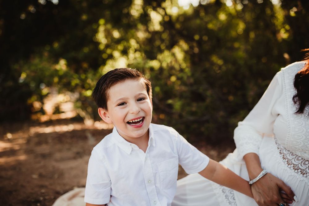 A joyful young boy in a white shirt laughs brightly outdoors, surrounded by lush green trees in warm sunlight near redlands ca