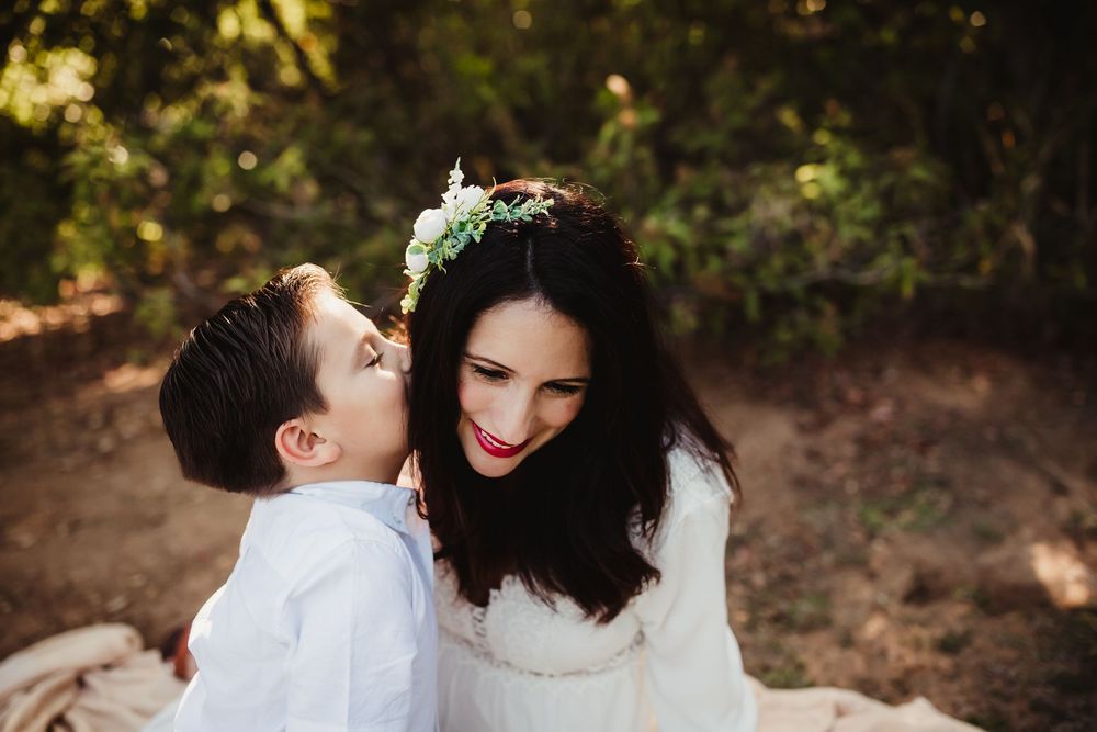 A young boy in white shirt kisses a smiling woman wearing a floral crown outdoors.