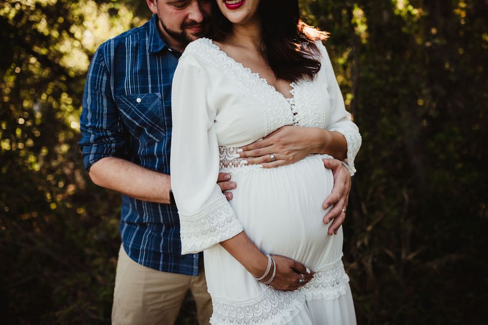 Couple embracing outdoors, pregnant woman in white lace dress with partner in blue plaid shirt cradling baby bump with heidi grace