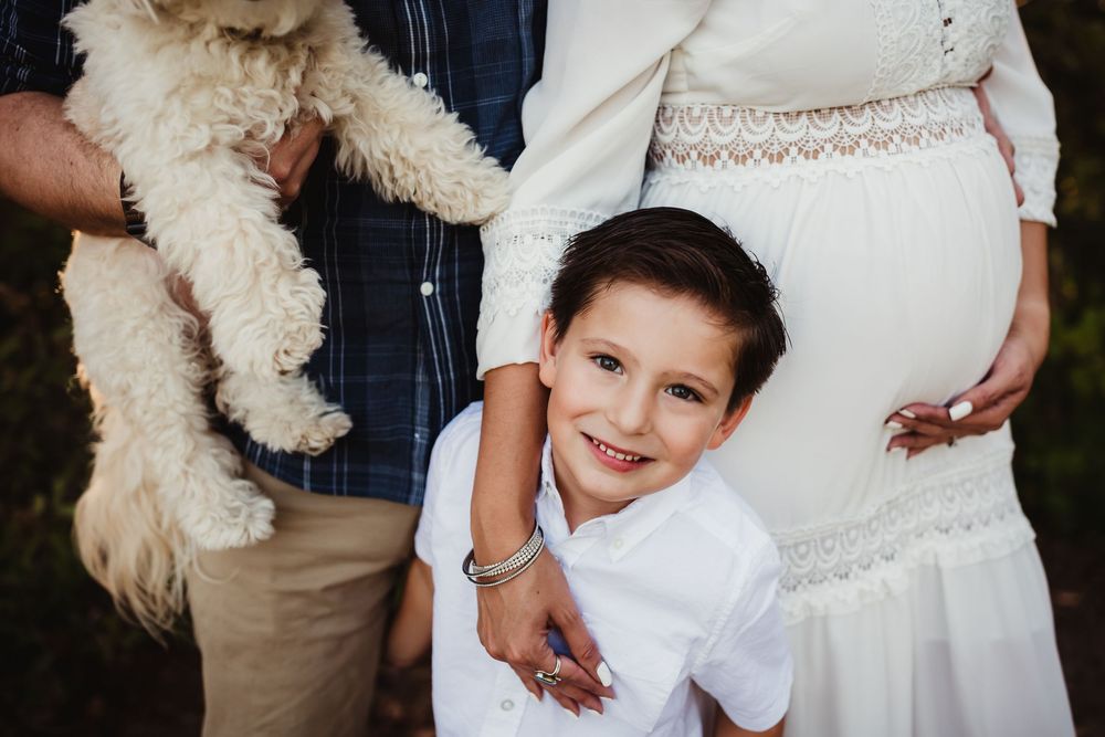 A smiling boy in white hugs a pregnant woman in a white dress, beside a man holding a fluffy dog outdoors.