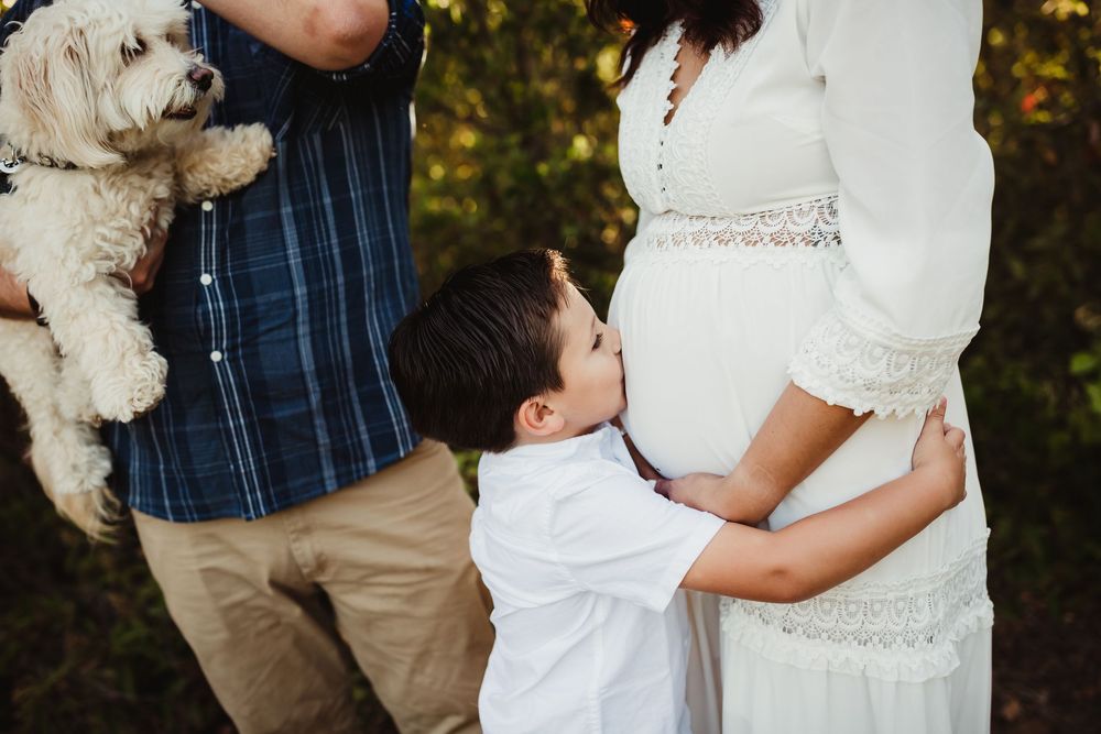 A young boy kisses his pregnant mother's belly during a family maternity photo shoot outdoors with a white dog.