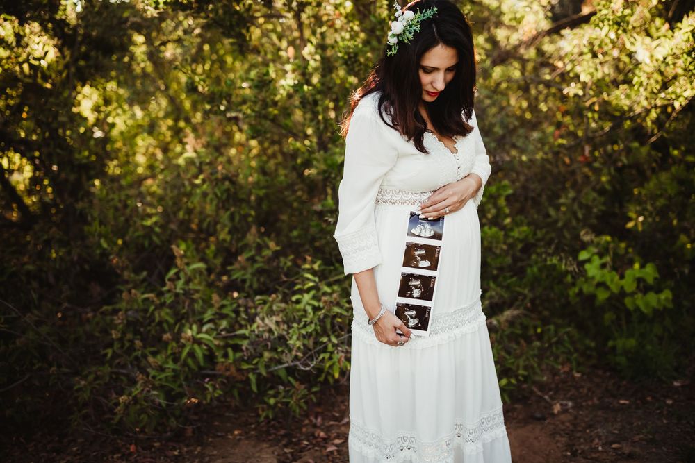 Pregnant woman in white dress holding ultrasound photos outdoors among green trees during maternity photo shoot with heidi grace